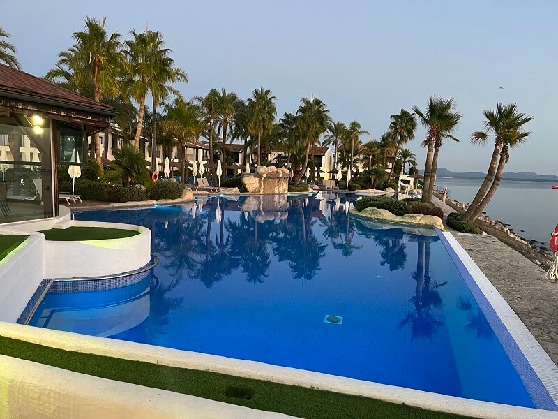 Port d‘Alcúdia - Pool eines Hotels mit Gartenlandschaft aus Palmen im Hintergrund