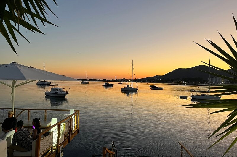 Port d‘Alcúdia - Blick auf die Bucht bei Dämmerung mit kleinen Schiffen, seitlich ins Bild ragenden Palmenblättern und dem Ausschnitt einer Chill-Terrasse mit weißem aufgespannten Sonnenschirm und sitzenden Menschen